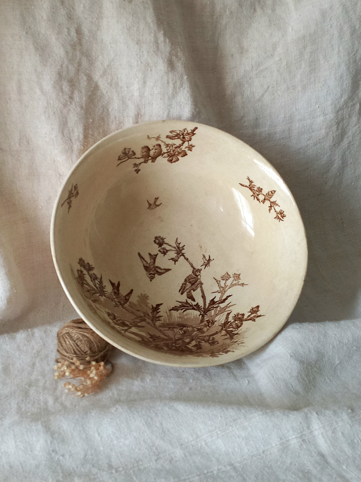 An off-white ceramic bowl with a brown sepia floral and bird pattern, resting on a textured white fabric background next to a small decorative object.