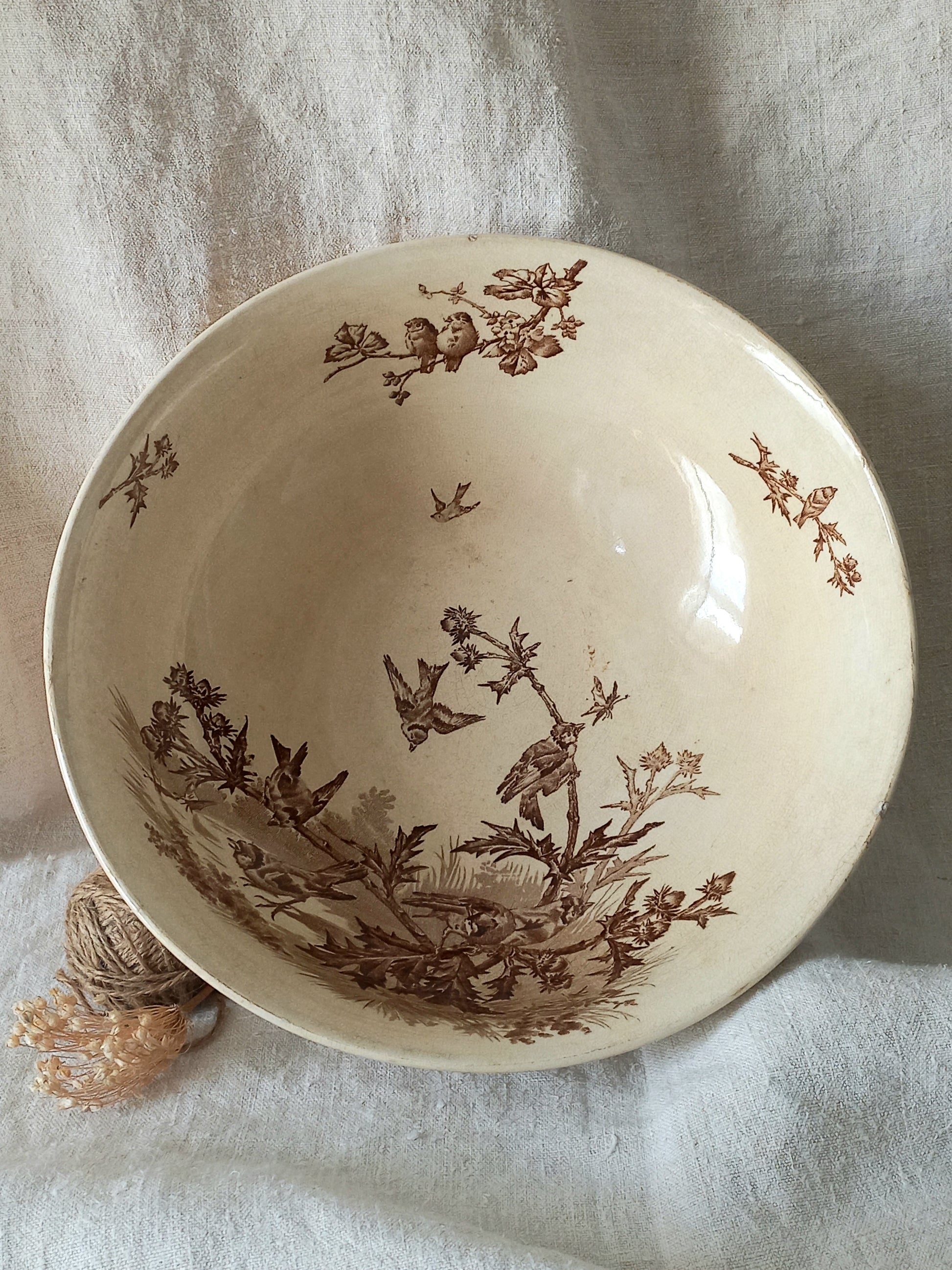 An off-white ceramic bowl with a brown sepia transfer print of birds, flowers, and foliage, displayed on a textured white fabric background.