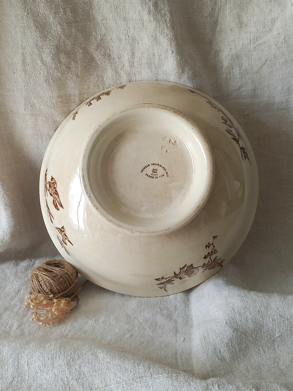 An antique cream-colored ceramic bowl with brown floral illustrations, shown upside down on a draped white linen fabric with a small ball of twine in the foreground.