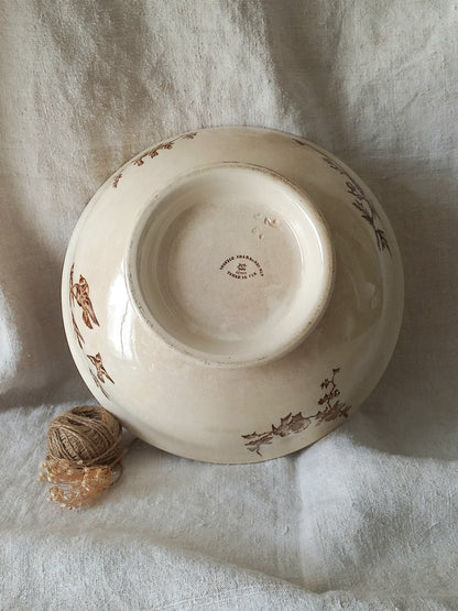 An antique cream-colored ceramic bowl with brown floral illustrations, shown upside down on a draped white linen fabric with a small ball of twine in the foreground.