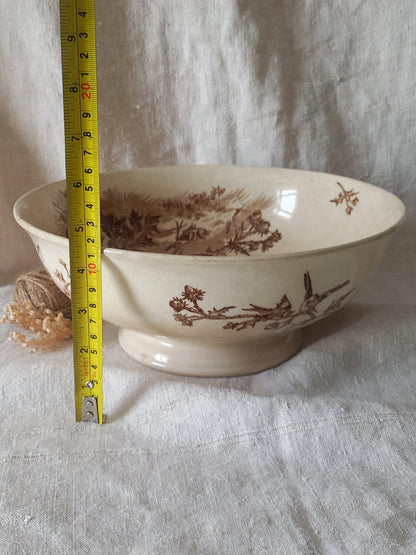 An off-white ceramic bowl with a brown sepia landscape and bird pattern, shown with a yellow tape measure standing next to it on a white fabric background.