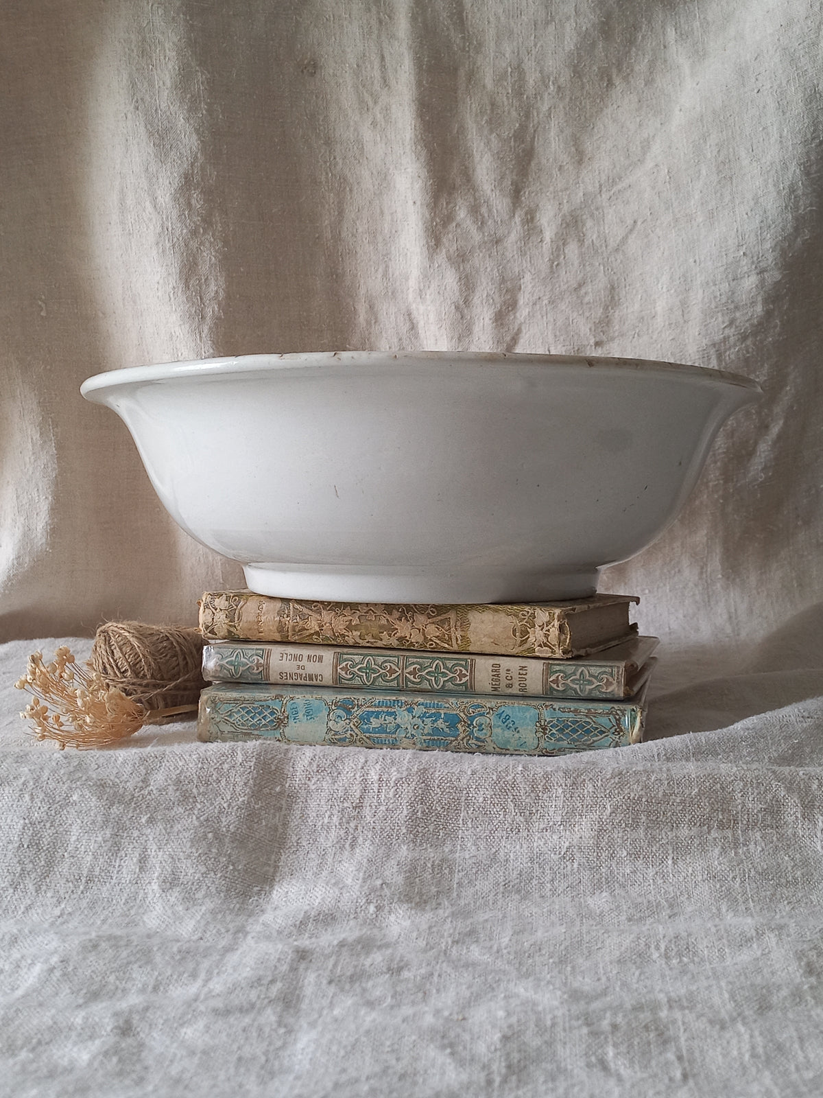 A large white ceramic bowl sits on top of a stack of vintage patterned books, with a small ball of twine and dried flowers in the foreground, set against a draped linen fabric backdrop.