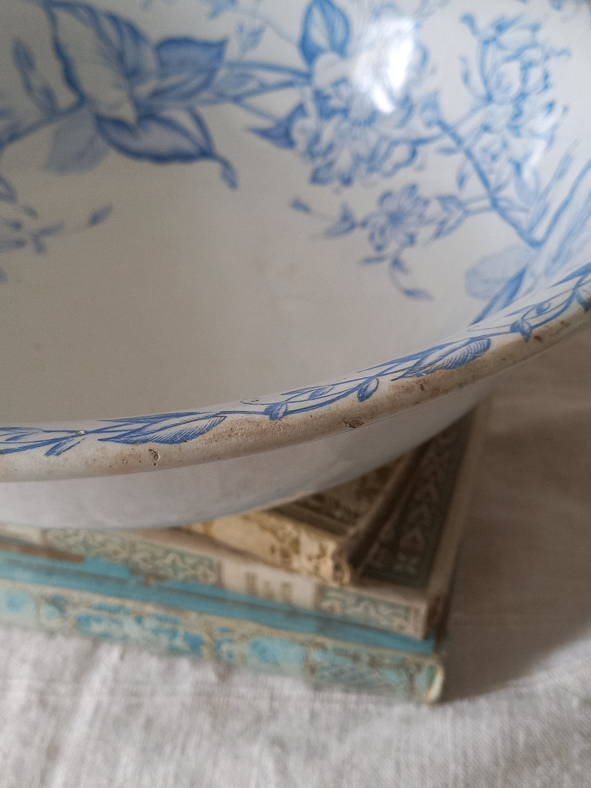 A close-up of a white ceramic bowl with a blue floral pattern, resting on top of vintage books on a white textured fabric surface.