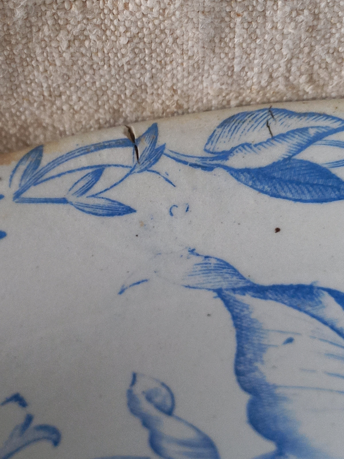Close-up of a blue and white patterned ceramic plate with a visible crack on the surface, resting on a textured beige fabric.