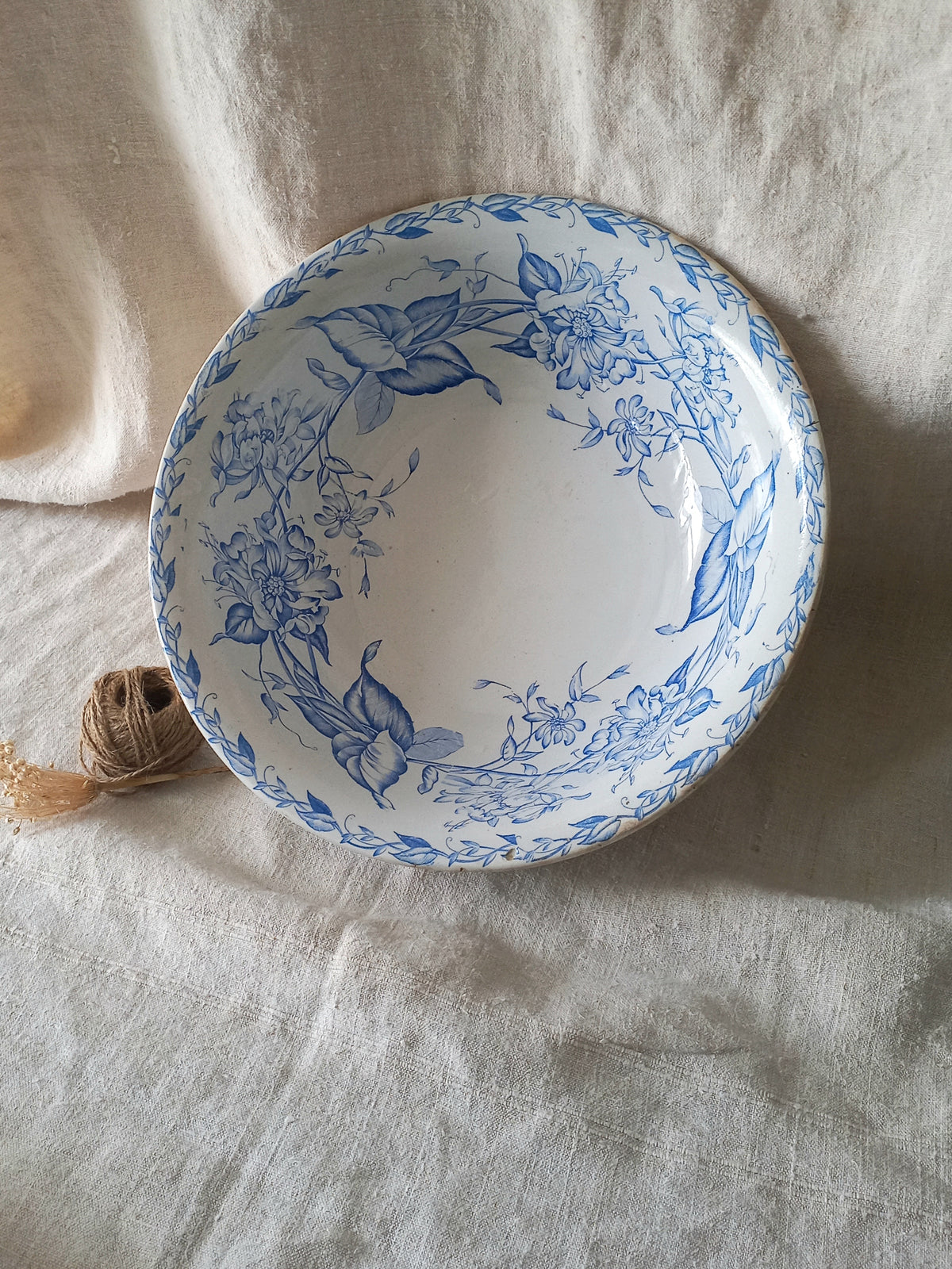 A round white ceramic bowl with a blue floral pattern, styled on a textured beige linen cloth next to a small ball of twine.