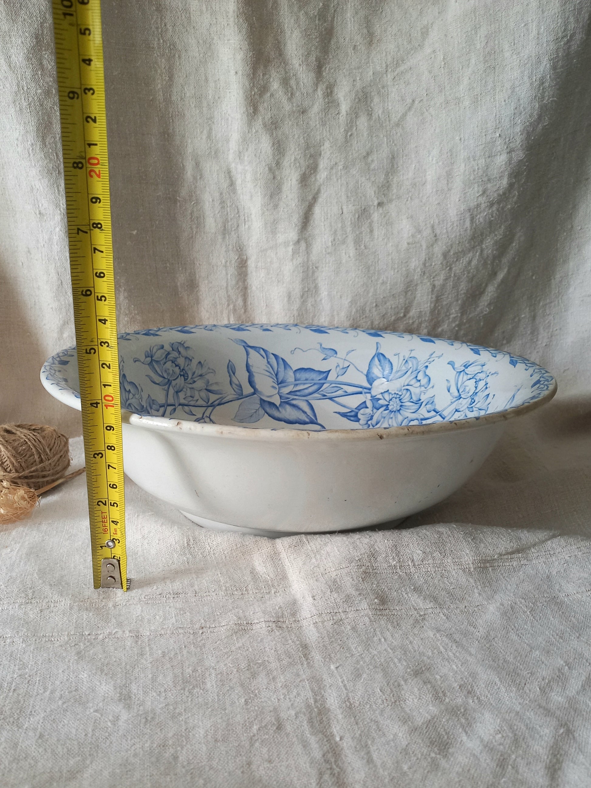 A white ceramic bowl with a blue floral pattern inside, shown next to a yellow tape measure for scale against a draped white linen background.