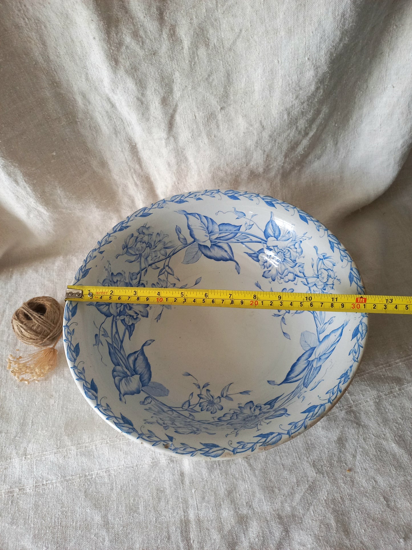 A blue and white porcelain bowl with a floral pattern, shown with a yellow tape measure across the rim to indicate its diameter, set against a draped white linen background.