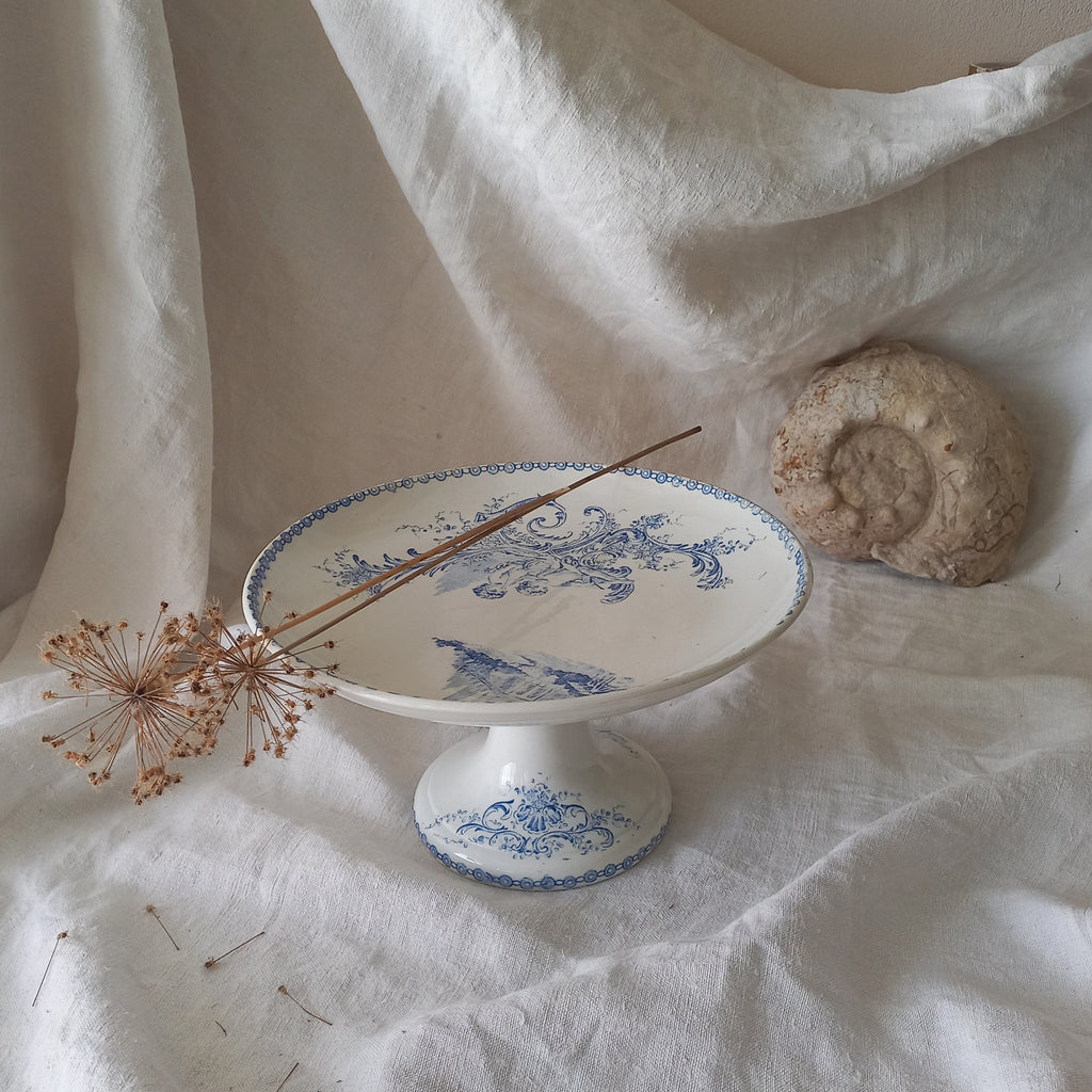 An attractive French antique footed ironstone cake plate with a blue transfer pattern depicting architecture, cherubs, and flowers; a very pretty and delicate pattern.

Original antique ironstone pieces of this age and condition are becoming increasingly difficult to source to this a rare treat to discover.
