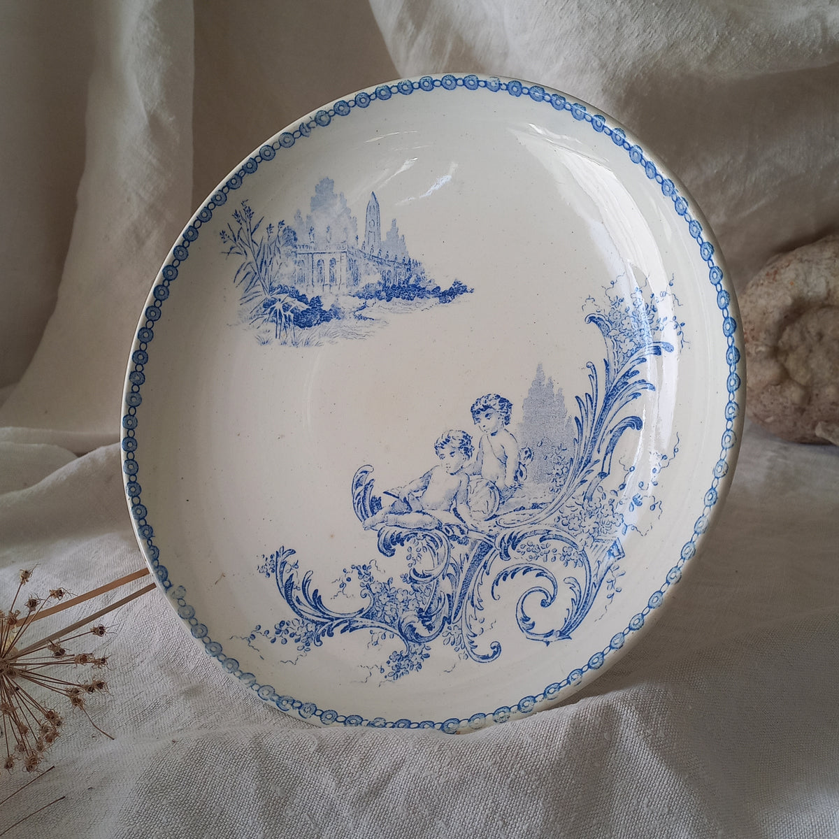 An attractive French antique footed ironstone cake plate with a blue transfer pattern depicting architecture, cherubs, and flowers; a very pretty and delicate pattern.

Original antique ironstone pieces of this age and condition are becoming increasingly difficult to source to this a rare treat to discover.
