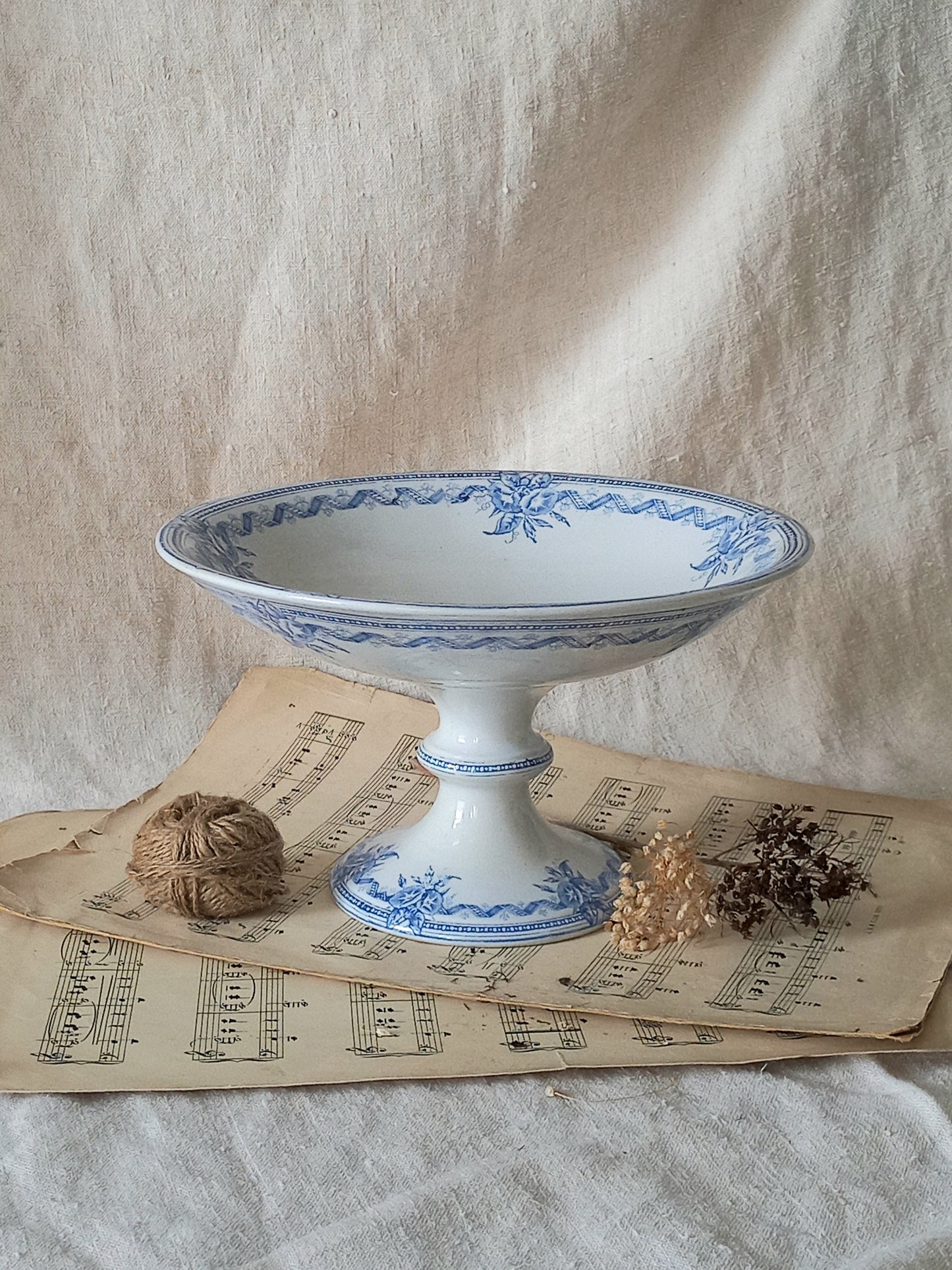 A white ceramic pedestal bowl with blue floral patterns, styled on vintage sheet music with twine and dried botanicals against a draped linen backdrop.