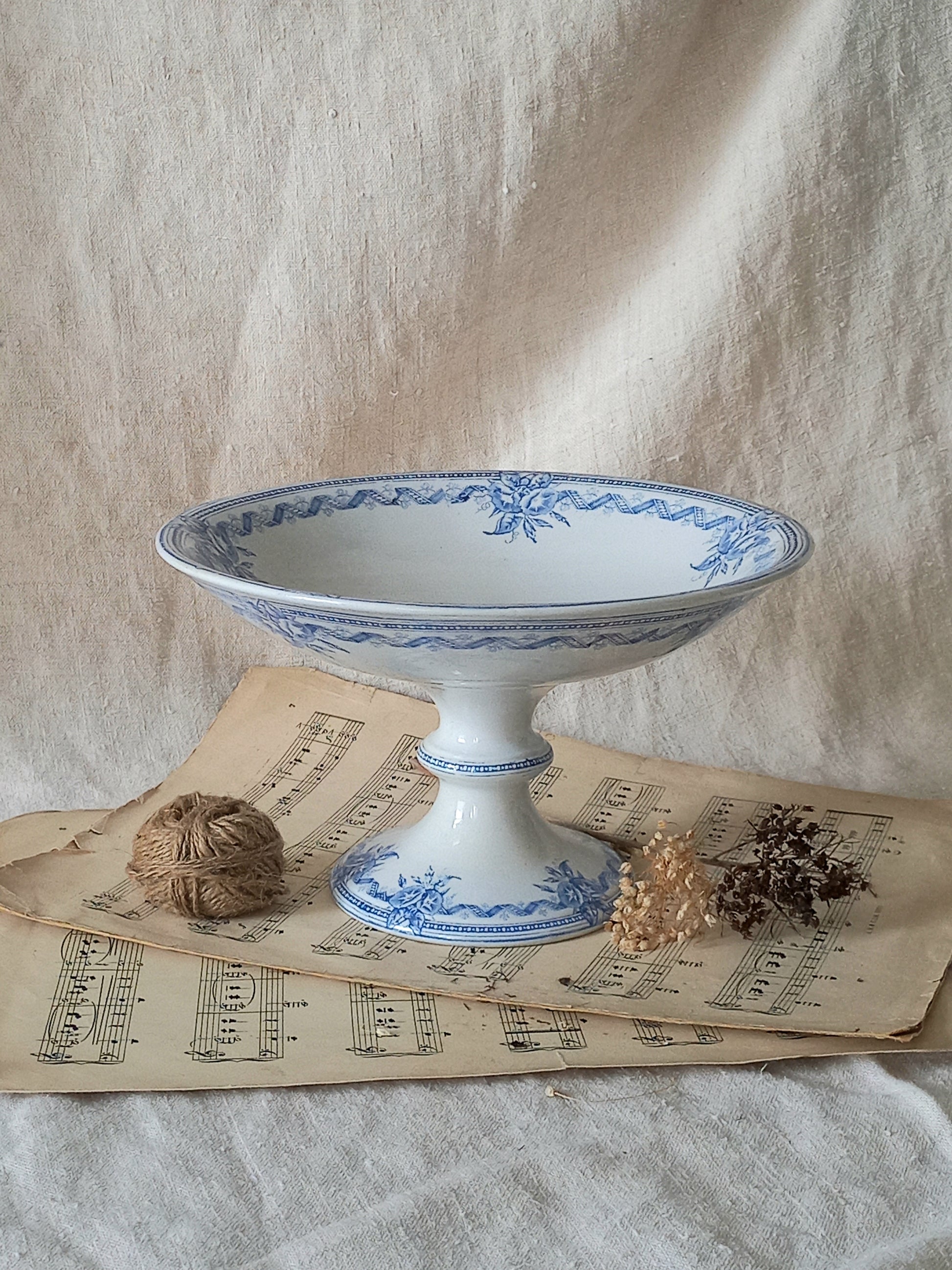 A white ceramic pedestal bowl with blue floral patterns, styled on vintage sheet music with twine and dried botanicals against a draped linen backdrop.