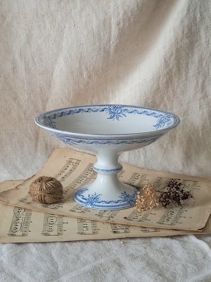 A white ceramic pedestal bowl with blue floral patterns, styled on vintage sheet music with twine and dried botanicals against a draped linen backdrop.