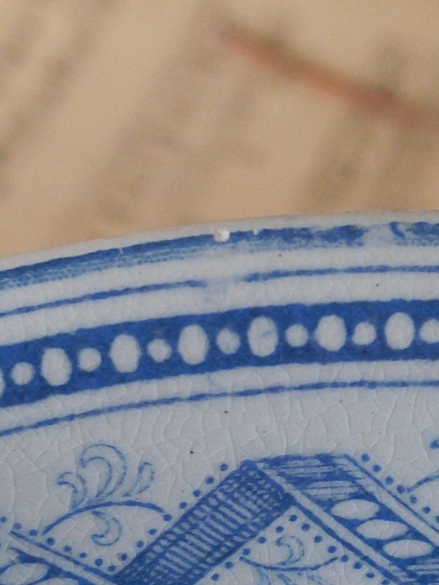 Close-up of a blue and white patterned ceramic surface with decorative bands and floral motifs, against a blurred beige background.