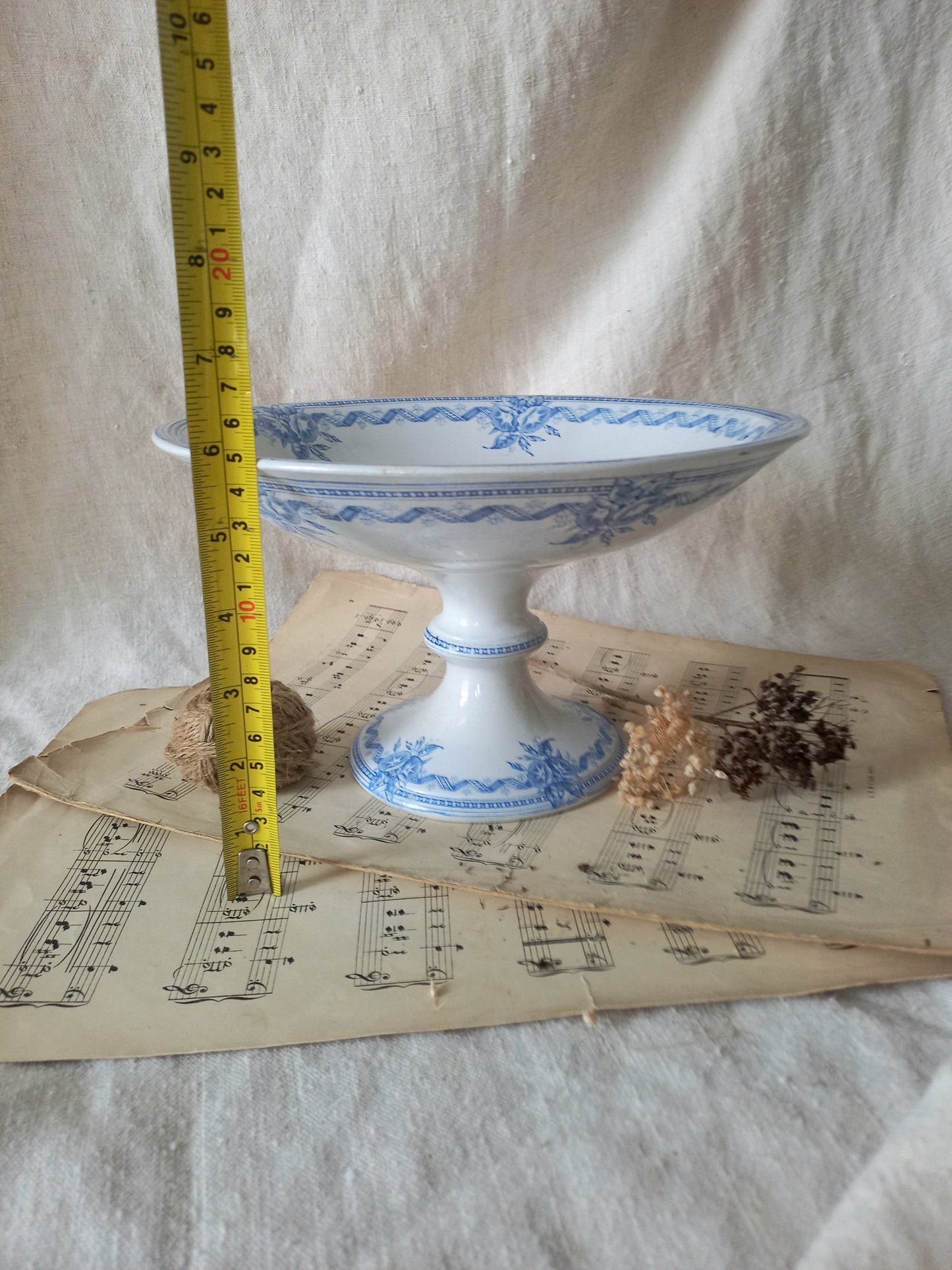 A white ironstone antique pedestal bowl with blue floral patterns, shown with a yellow tape measure for scale on top of vintage sheet music and dried flowers against a draped white fabric background.