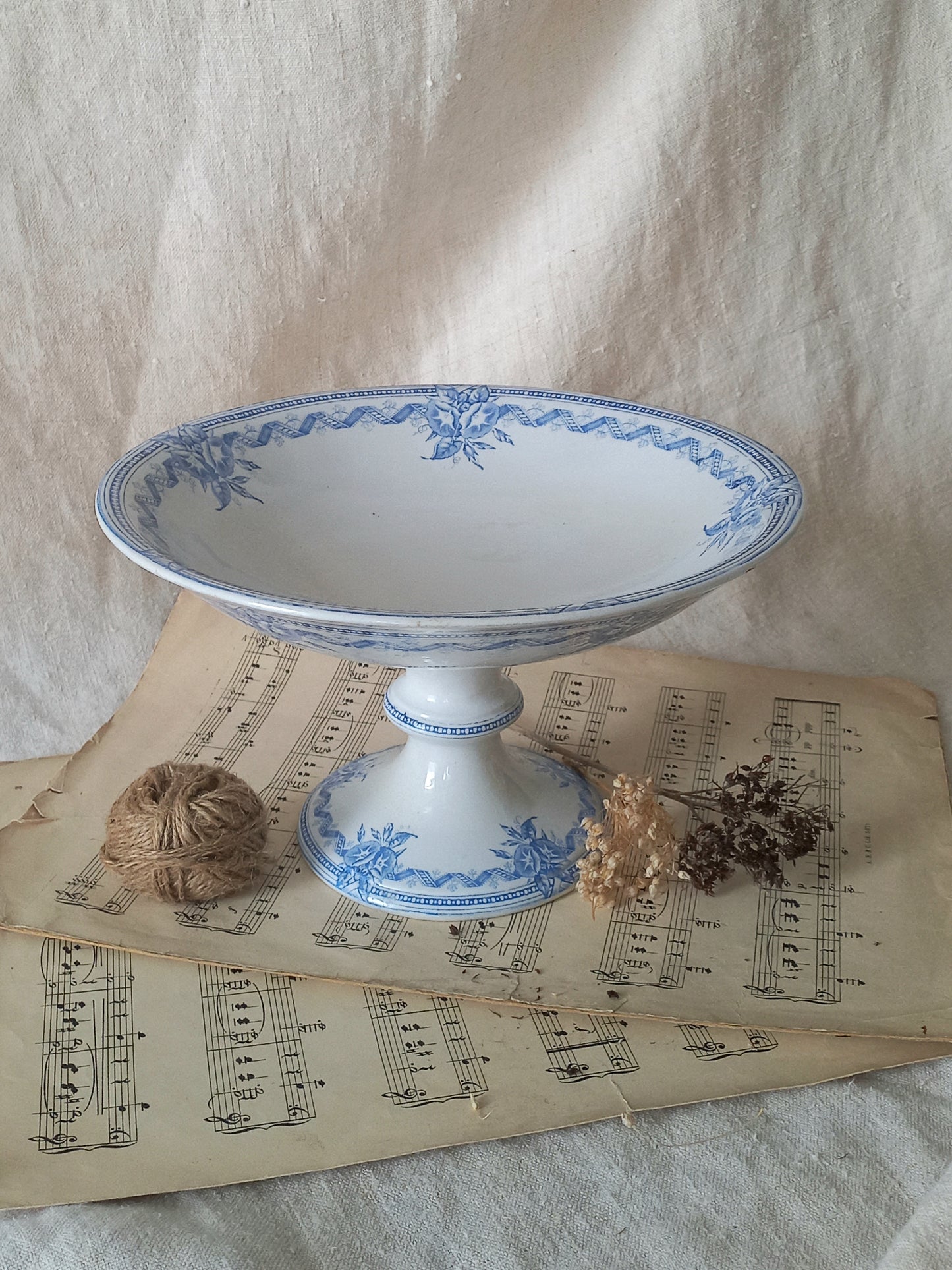 A white ceramic pedestal bowl with blue floral patterns, styled on vintage sheet music with twine and dried flowers against a draped fabric backdrop.