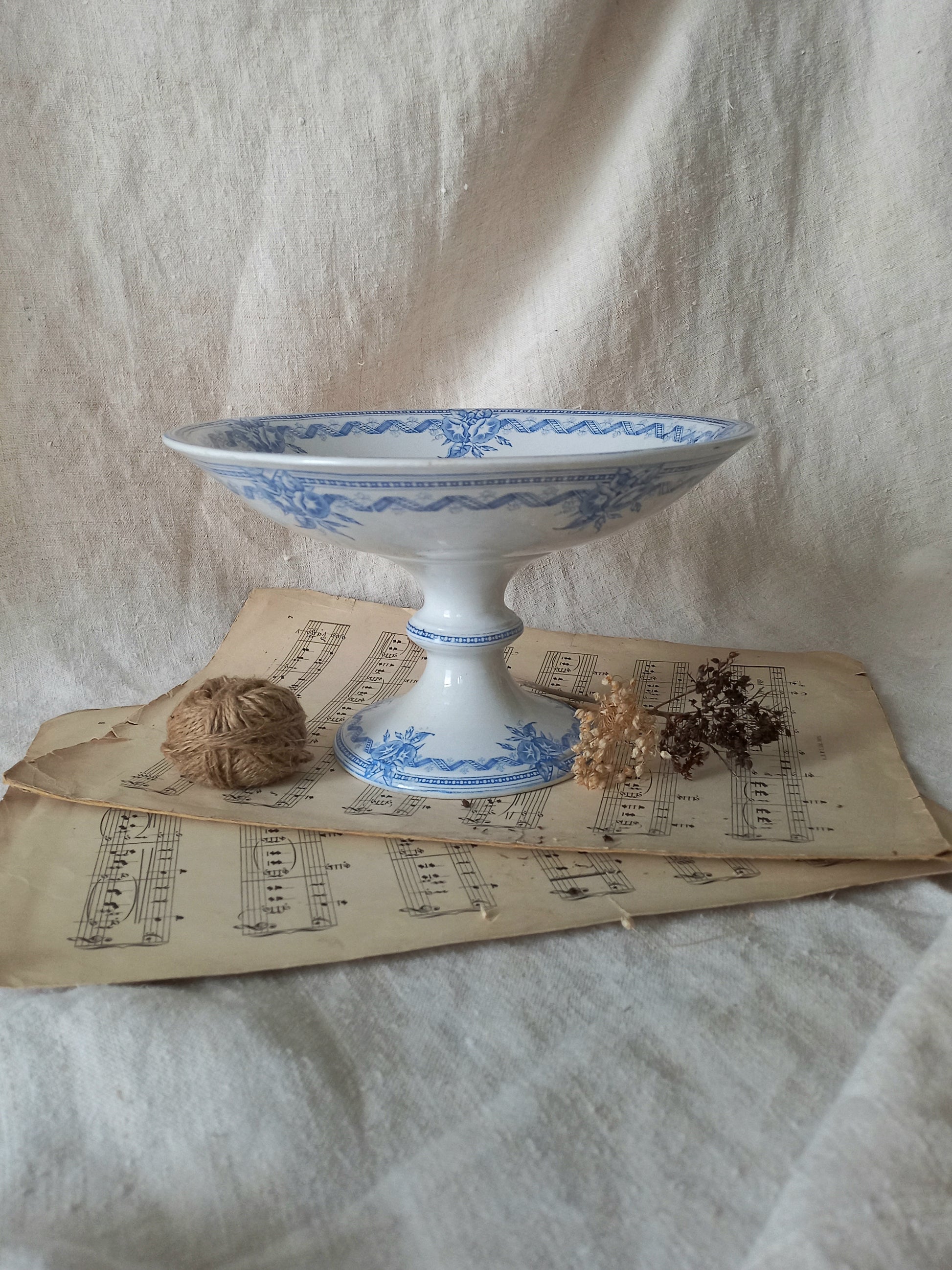 A white ceramic pedestal bowl with blue floral patterns, styled on vintage sheet music with dried flowers and a small brown decorative object against a draped cream fabric backdrop.