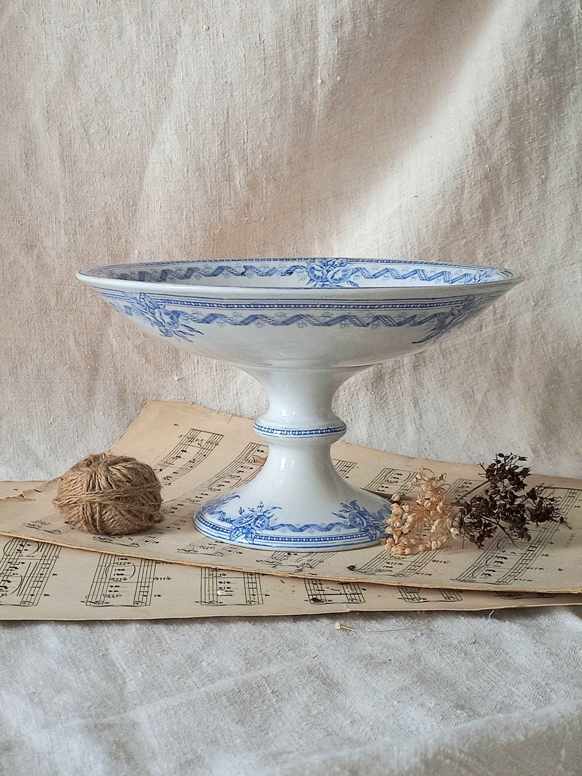 A white ironstone antique pedestal bowl with blue floral and geometric patterns, styled on vintage sheet music with a twine ball and dried flowers against a draped beige fabric backdrop.