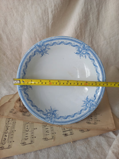 A round white ceramic footed bowl with a blue floral border pattern, shown with a yellow tape measure across the rim to indicate size, resting on vintage sheet music against a beige fabric backdrop.