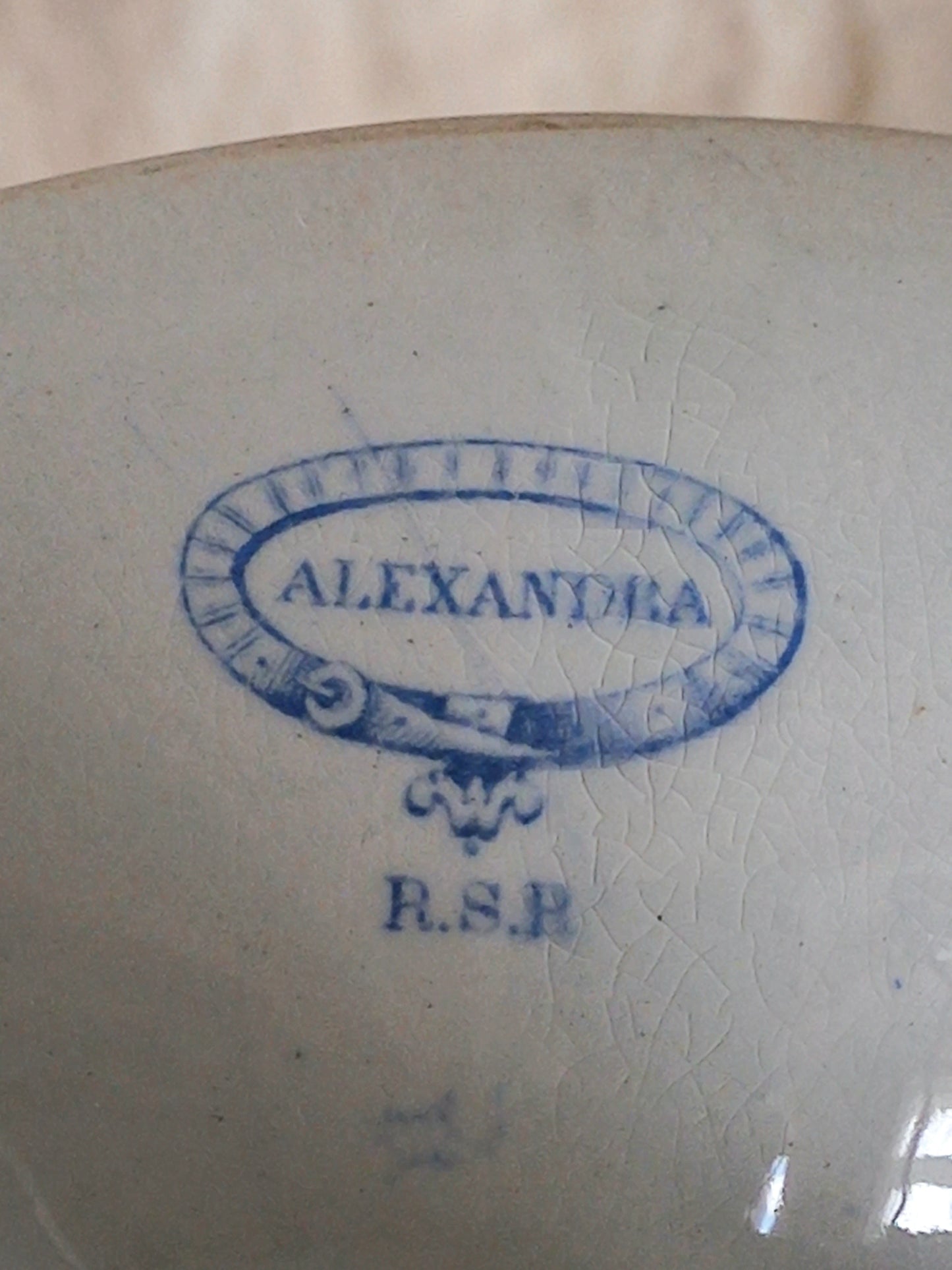 Close-up of the base of a white footed bowl with a blue printed monogram or brand mark reading 'ALEXANDRA' 