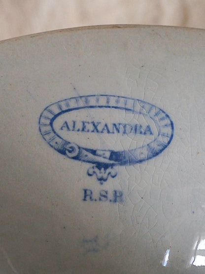 Close-up of the base of a white footed bowl with a blue printed monogram or brand mark reading 'ALEXANDRA' 