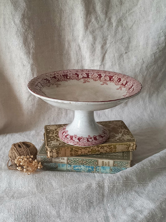 A vintage white ceramic pedestal stand with a pink floral patterned rim, displayed on a stack of antique books next to a ball of twine with small beads, set against a draped off-white linen backdrop.