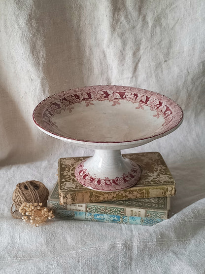 An antique ceramic pedestal plate with a pink /burgandy and white floral border, displayed on top of stacked antique books with a small ball of twine nearby, set against a draped white fabric backdrop.