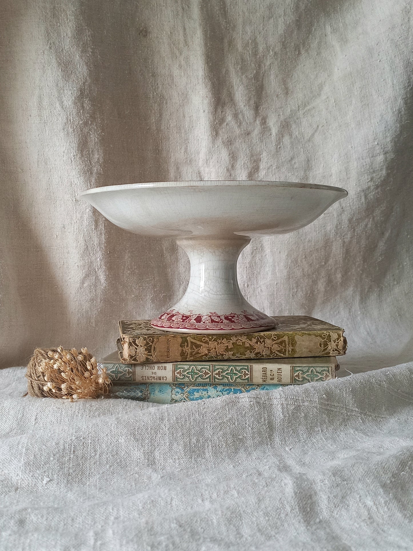 A white ceramic pedestal bowl or cake stand displayed on top of three vintage books, with a small gold-toned decorative object beside them, set against a draped off-white fabric backdrop.