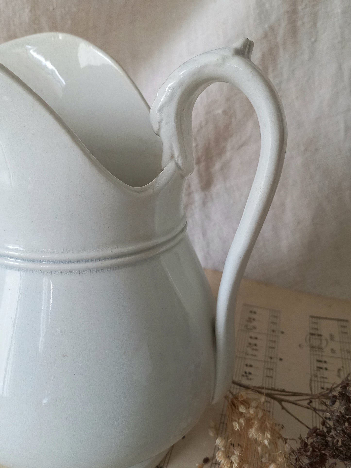 A white ceramic pitcher with a curved handle and spout, shown close-up against a soft beige fabric background with dried flowers and a vintage paper sheet nearby.