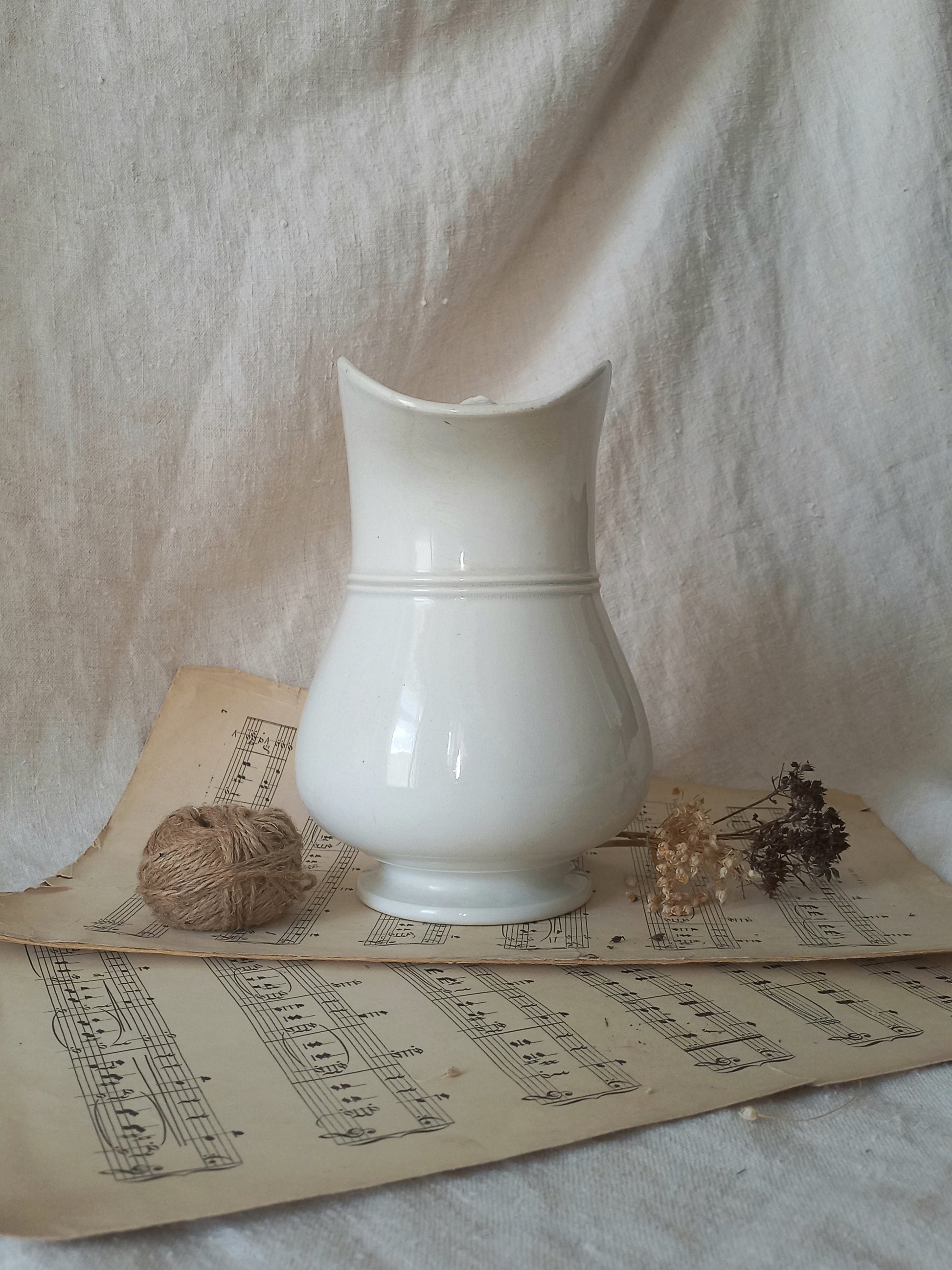 A glossy white ceramic pitcher with a curved spout, styled on vintage sheet music with a ball of twine and dried flowers against a draped linen backdrop.