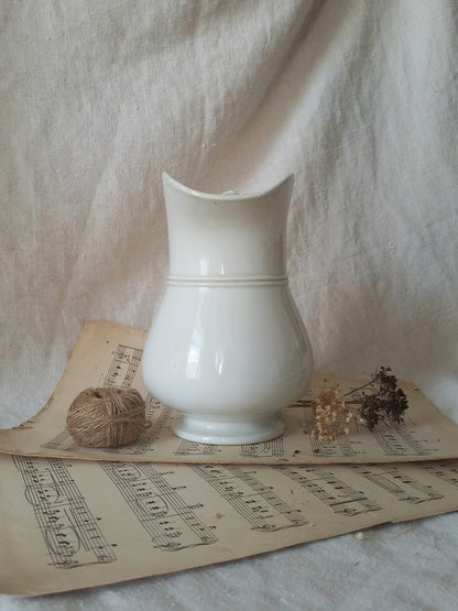 A glossy white ceramic pitcher with a curved spout, styled on vintage sheet music with a ball of twine and dried flowers against a draped linen backdrop.