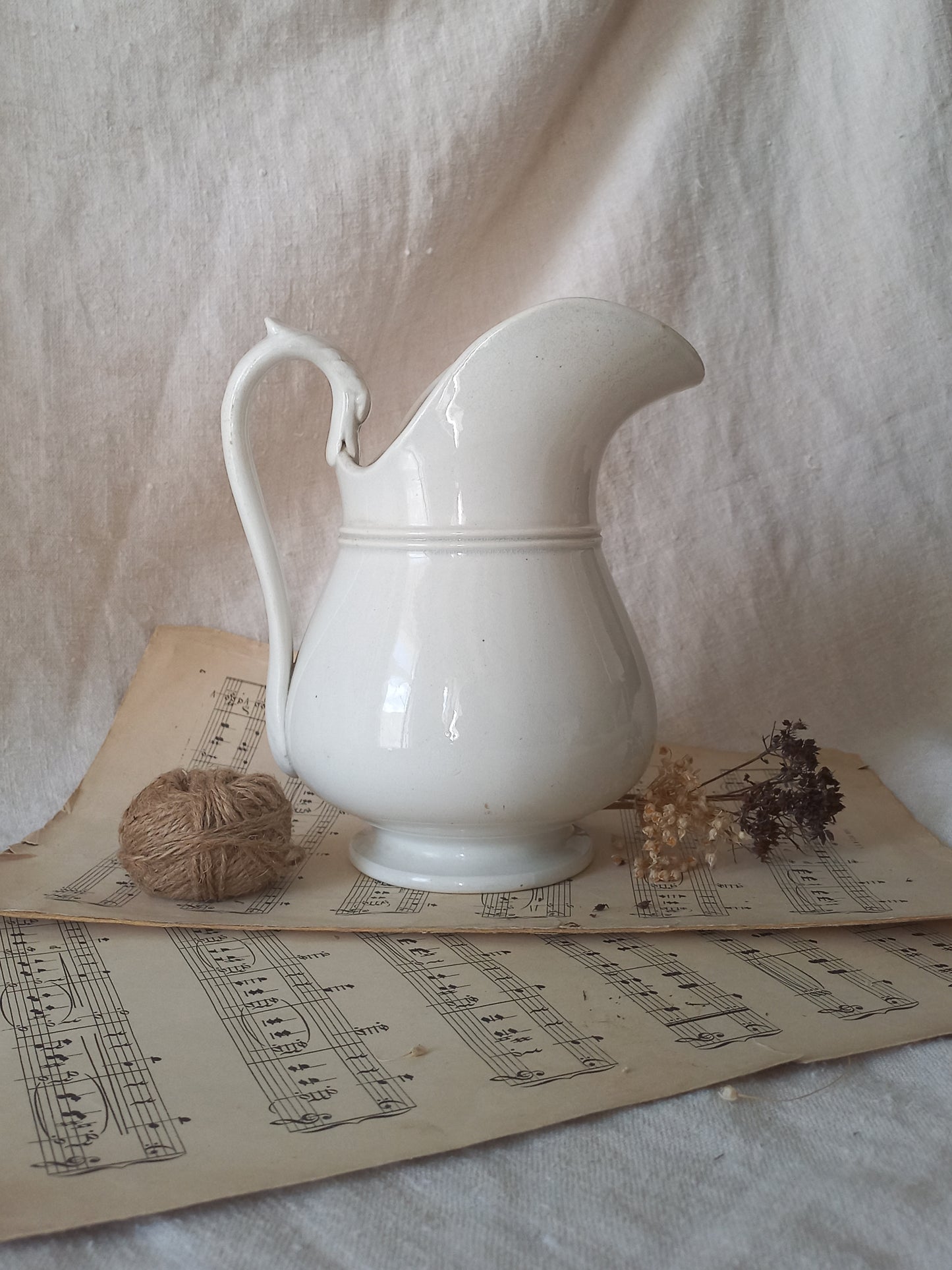 A white ceramic pitcher with a curved handle and spout, styled on vintage sheet music with a ball of twine and dried flowers against a draped fabric backdrop.