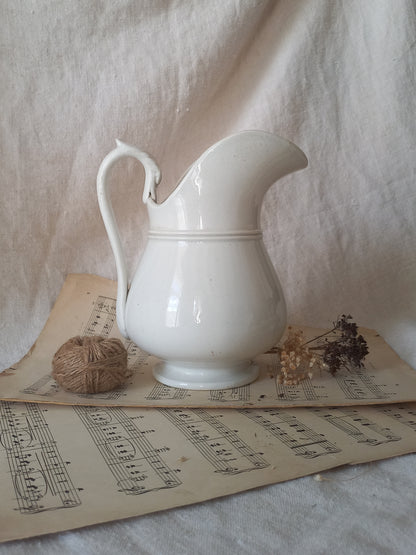 A white ceramic pitcher with a curved handle and spout, styled on vintage sheet music with a ball of twine and dried flowers against a draped fabric backdrop.