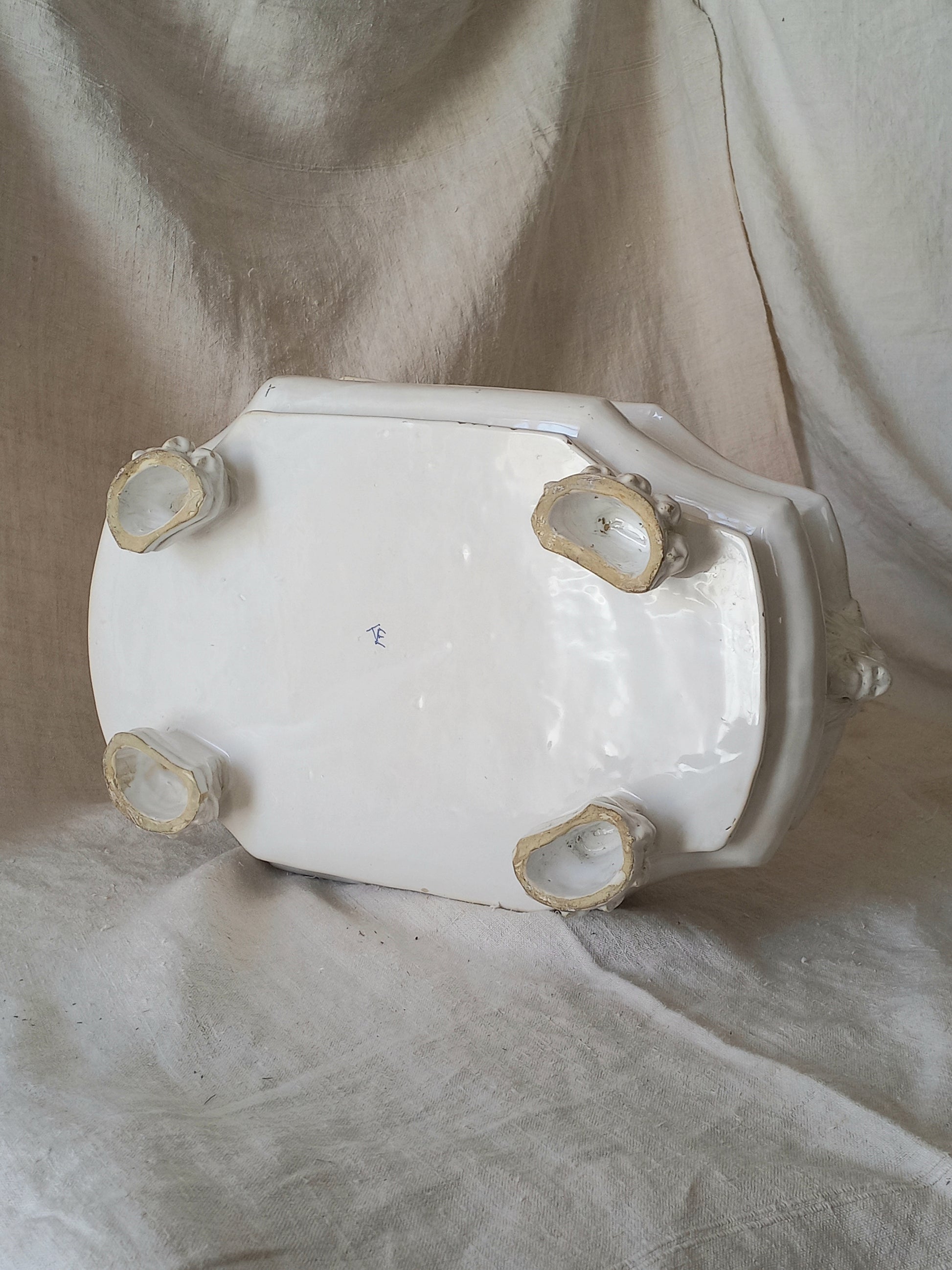 Underneath an antique white ceramic planter with a lion head relief on the front and claw-footed base, set against a draped beige fabric background.