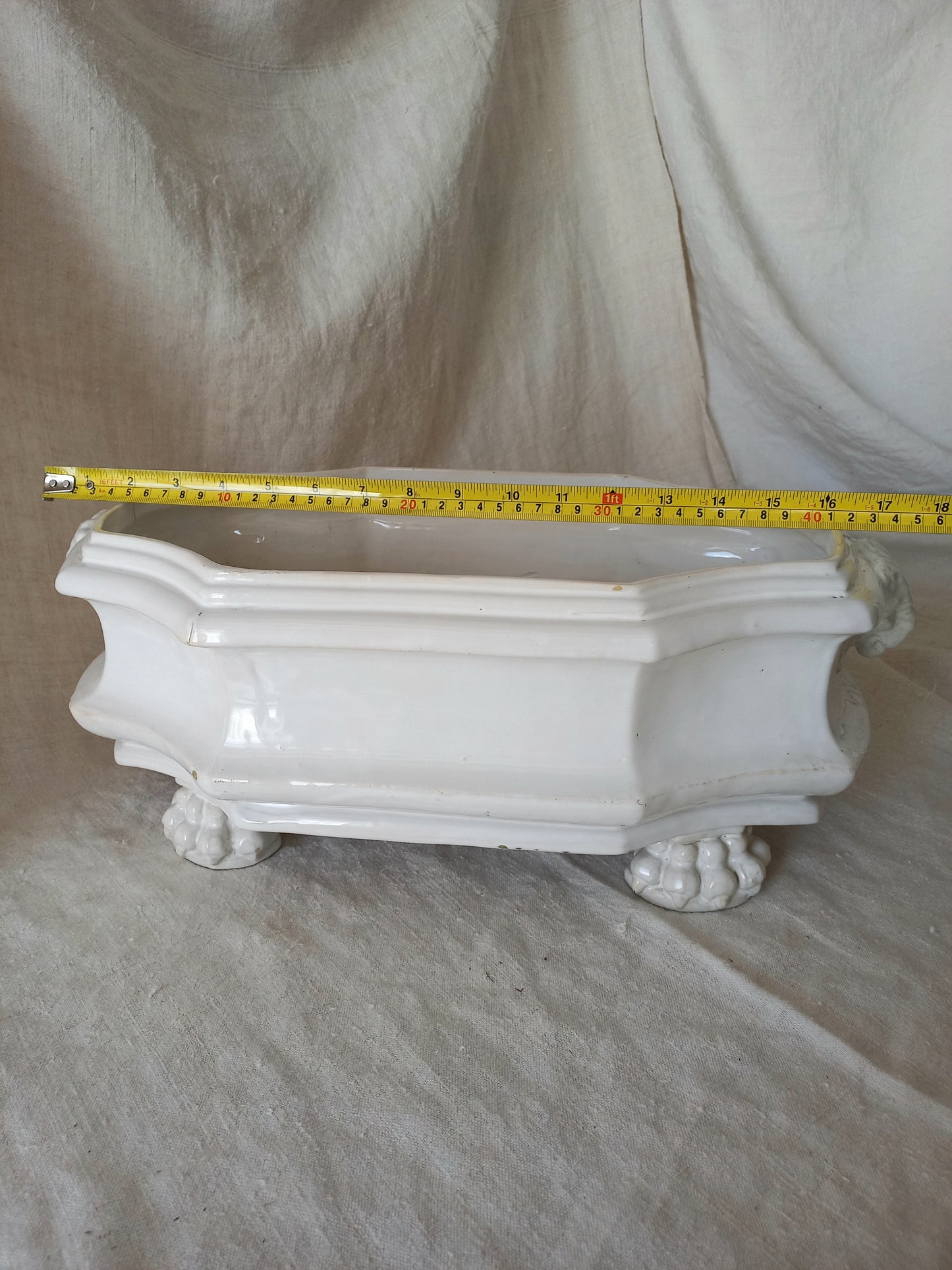 antique white ceramic planter with a lion head relief on the front and claw-footed base, set against a draped beige fabric background with tape measure for reference.