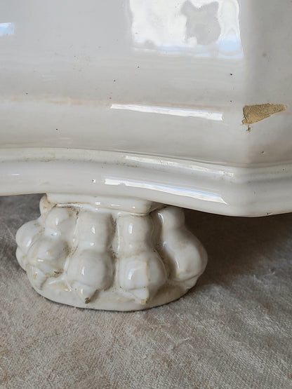 Close-up of an antique white ceramic planter with a decorative ribbed pedestal base, resting on a textured beige fabric surface.