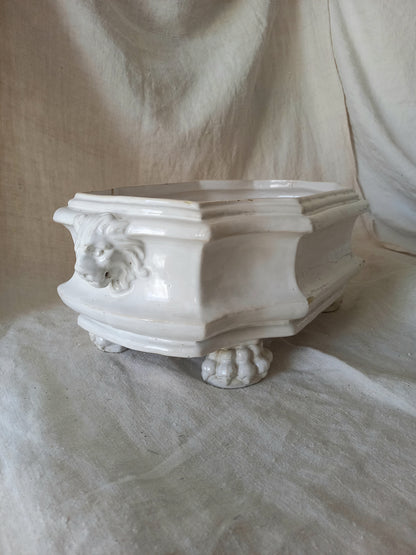 Close-up detail of antique white ceramic planter with a lion head relief on the front and claw-footed base, set against a draped beige fabric background.