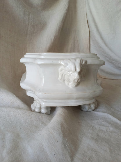 Close-up detail of antique white ceramic planter with a lion head relief on the front and claw-footed base, set against a draped beige fabric background.