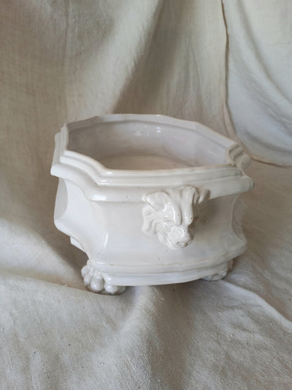 Close-up detail of antique white ceramic planter with a lion head relief on the front and claw-footed base, set against a draped beige fabric background.
