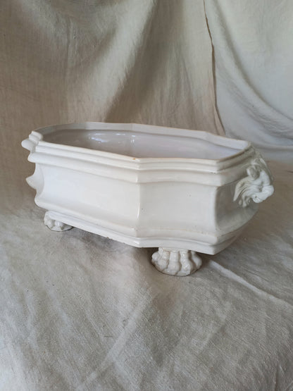 Close-up detail of antique white ceramic planter with a lion head relief on the front and claw-footed base, set against a draped beige fabric background.