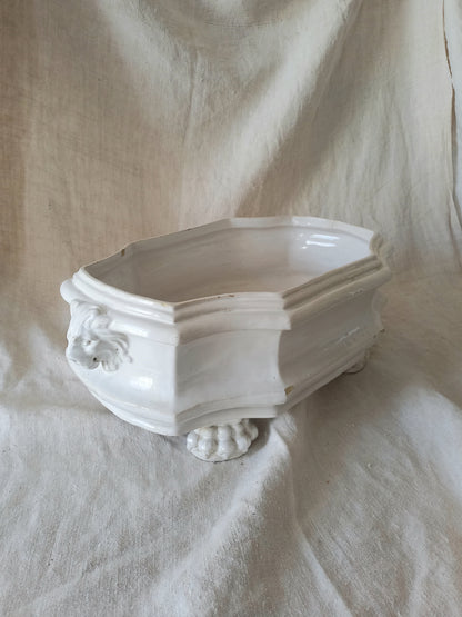 An antique white ceramic planter with a lion head relief on the front and claw-footed base, set against a draped beige fabric background.