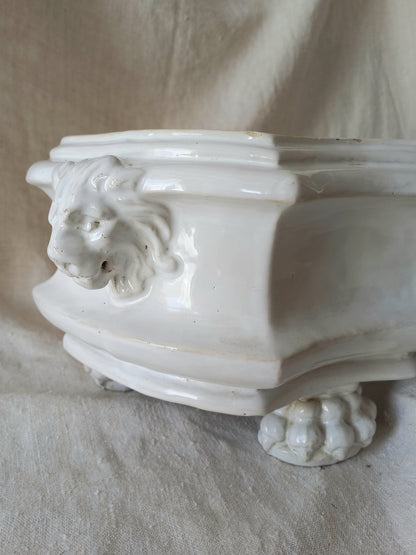 Close-up of an antique white ceramic planter with a lion head relief on the front and claw-footed base, set against a draped beige fabric background.