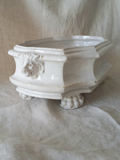 An antique white ceramic planter with a lion head relief on the front and claw-footed base, set against a draped beige fabric background.