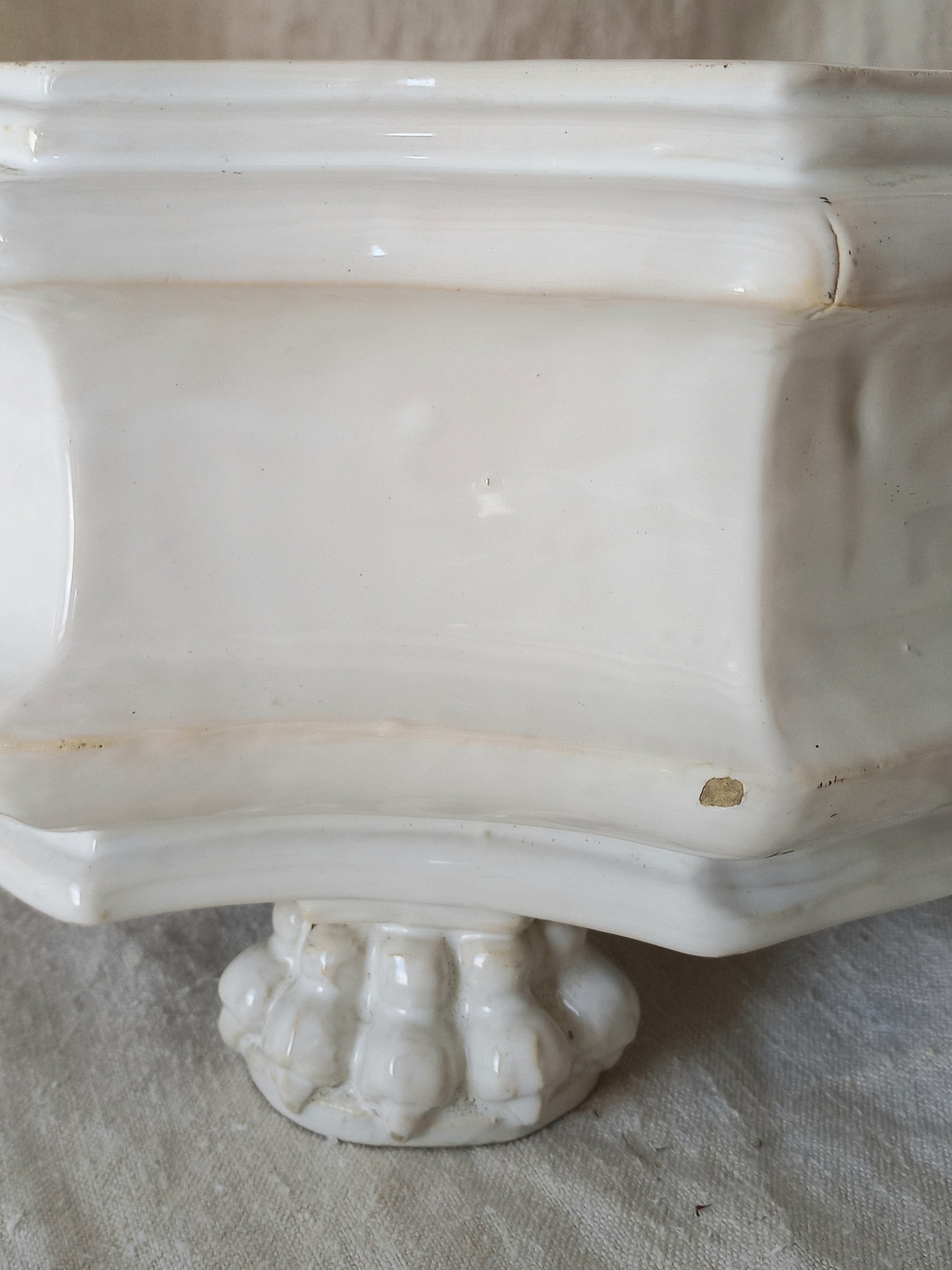Close-up detail of antique white ceramic planter with a lion head relief on the front and claw-footed base, set against a draped beige fabric background.