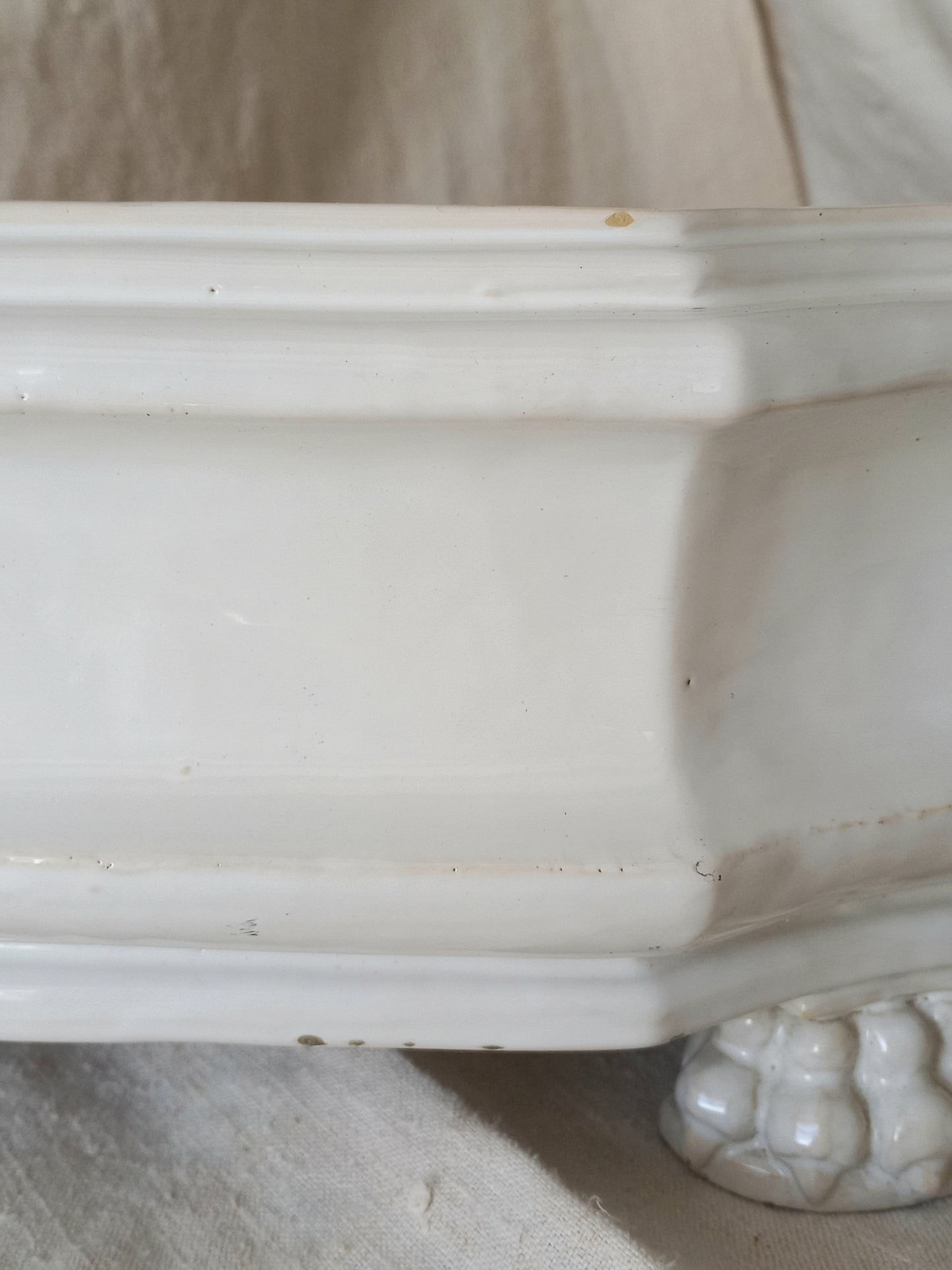 Close-up detail of antique white ceramic planter with a lion head relief on the front and claw-footed base, set against a draped beige fabric background.