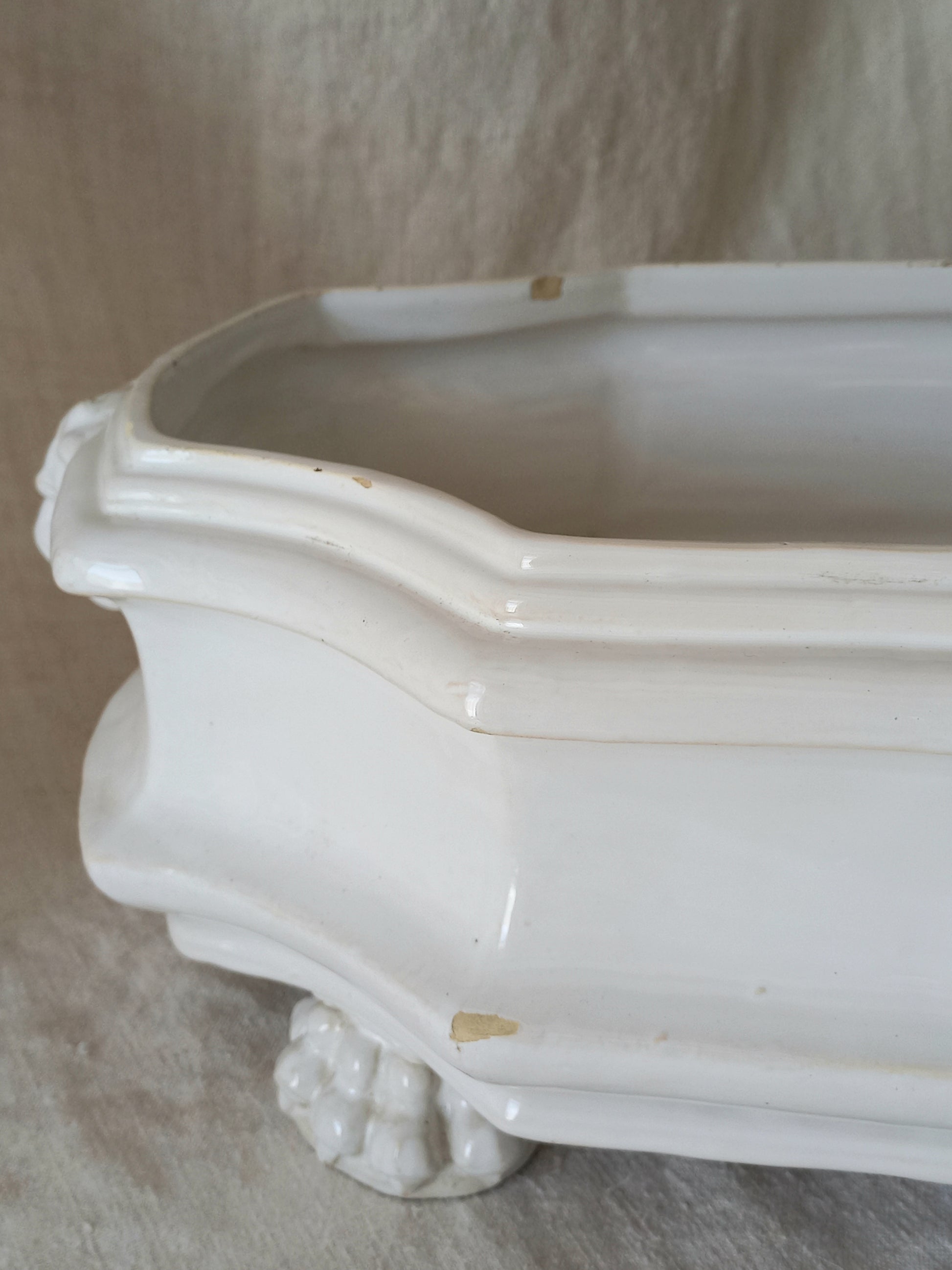 Close-up detail of antique white ceramic planter with a lion head relief on the front and claw-footed base, set against a draped beige fabric background.
