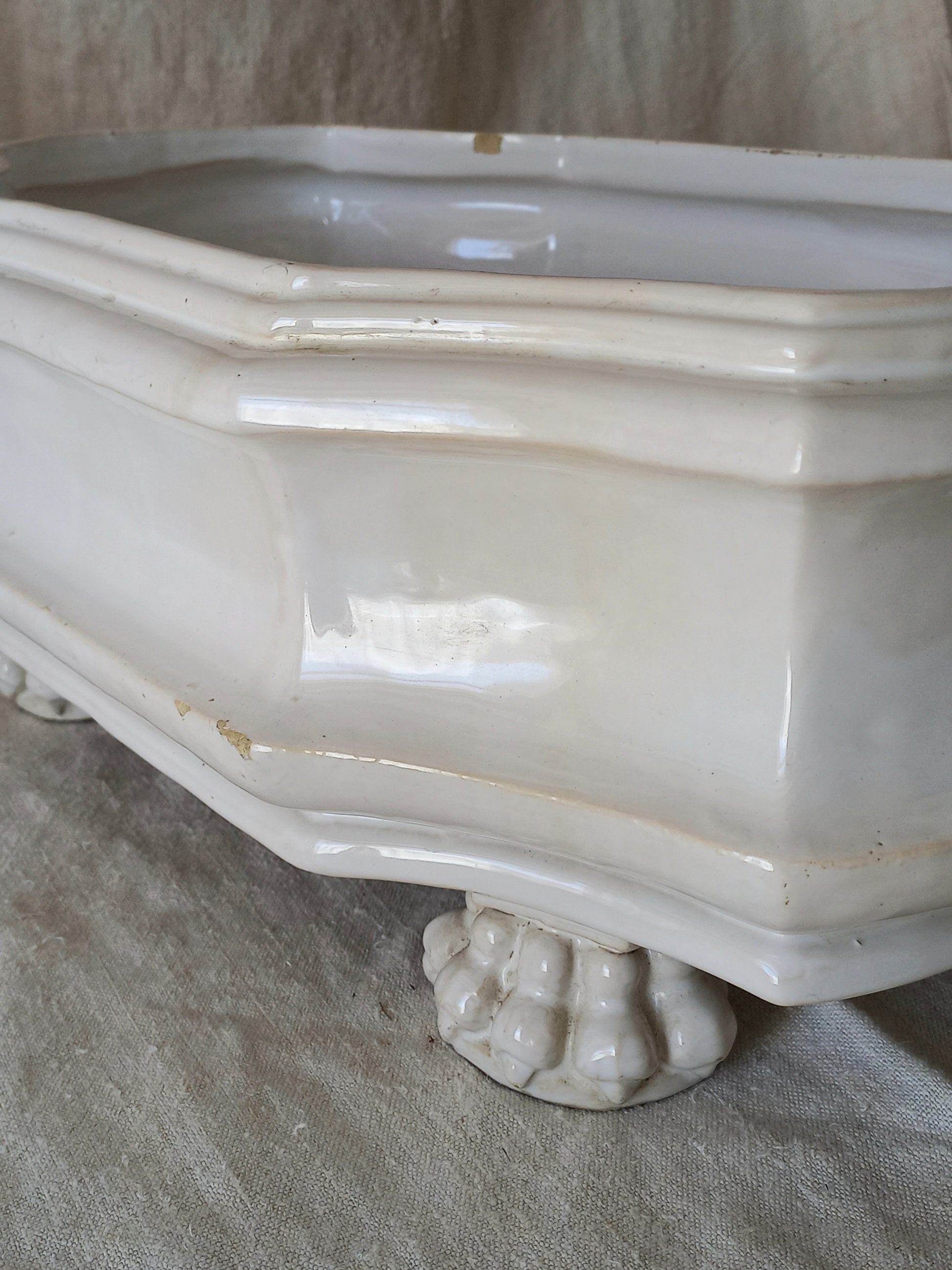 Close-up detail of antique white ceramic planter with a lion head relief on the front and claw-footed base, set against a draped beige fabric background.