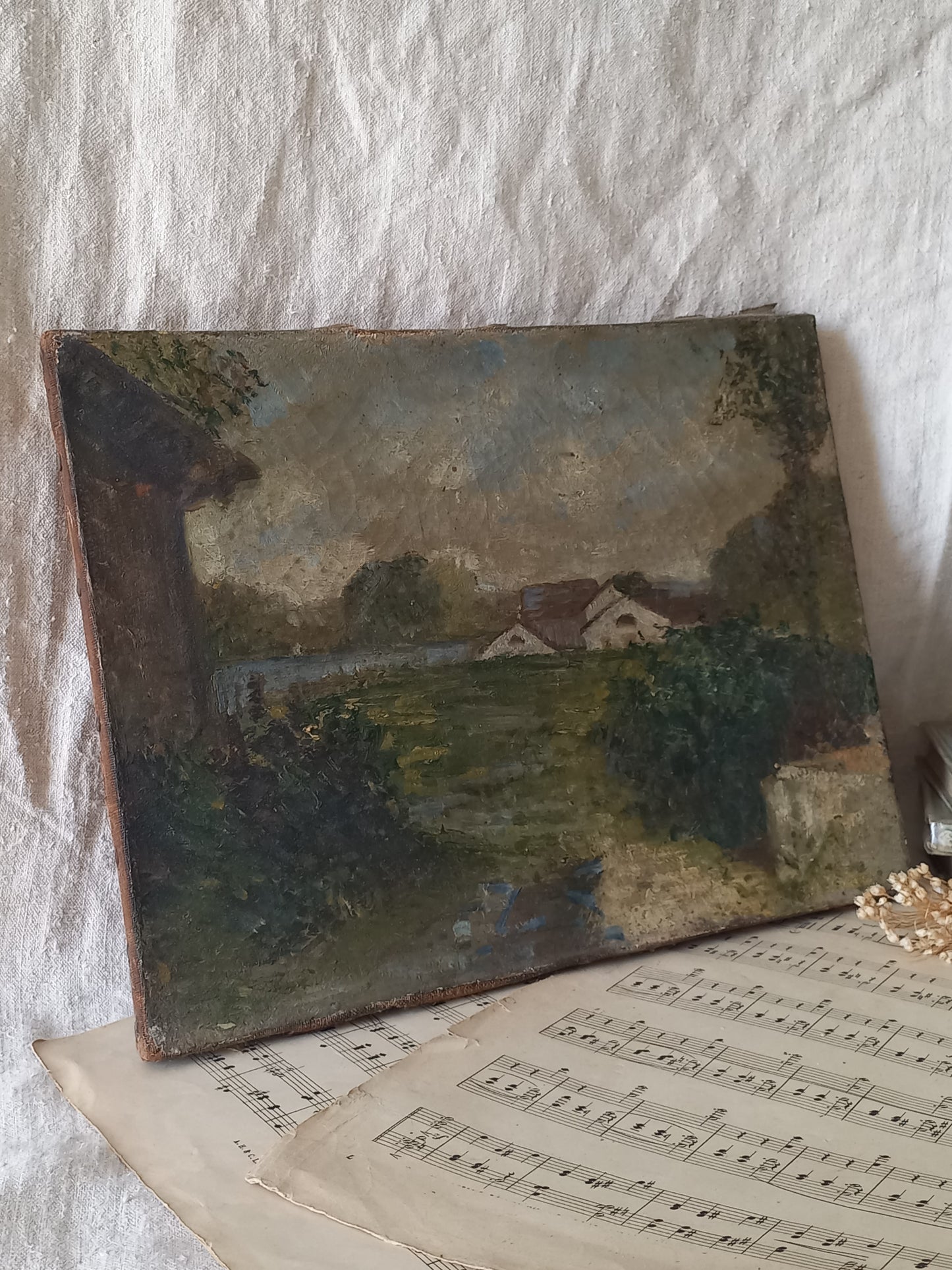 An antique landscape oil painting on canvas, depicting a rural scene with houses, trees, and a path, displayed on top of vintage sheet music against a textured white fabric background.