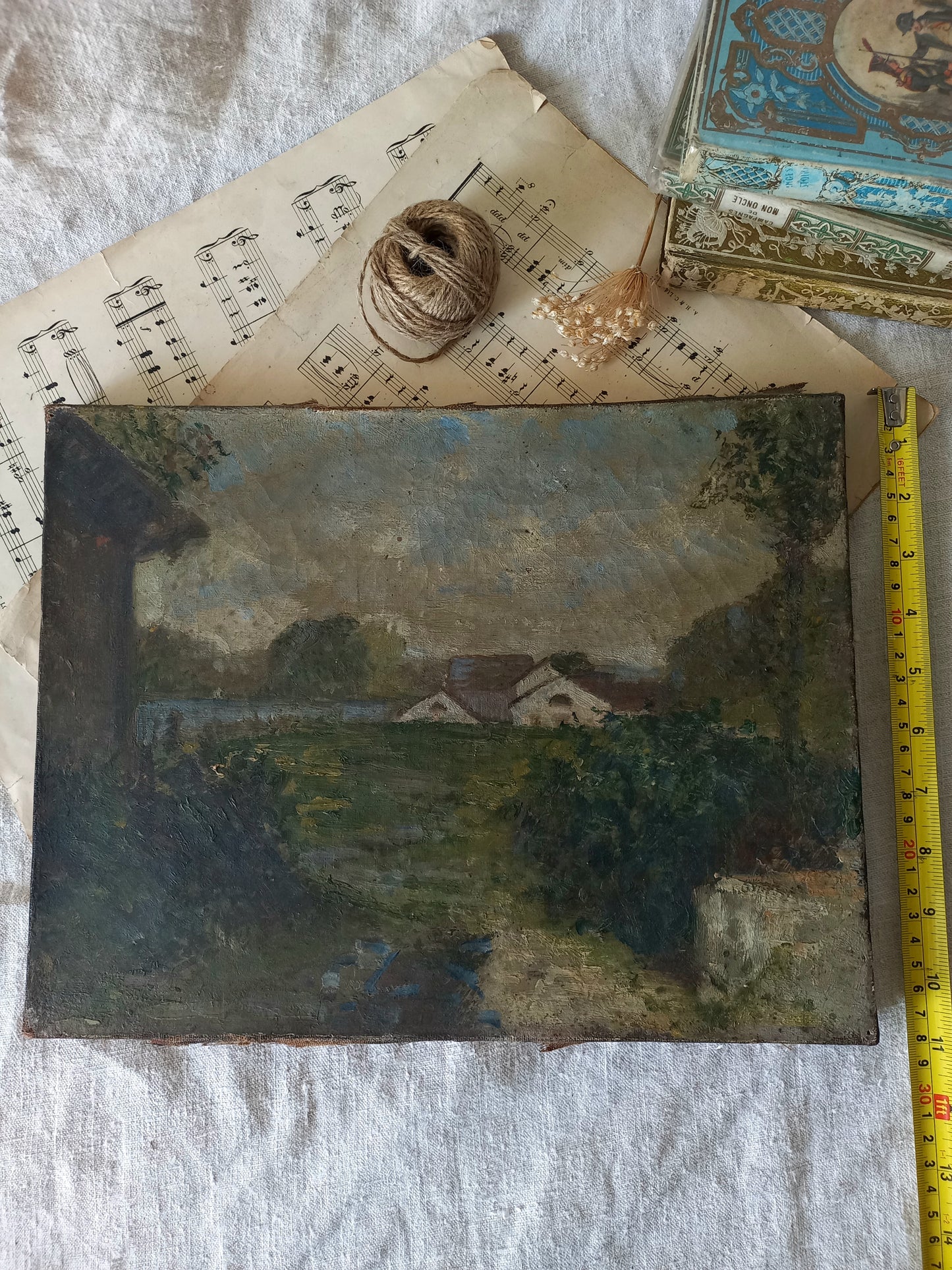 An antique oil landscape painting of a rural scene with houses, trees, and a path, displayed on a white fabric background with vintage sheet music, twine, dried flowers, and a yellow tape measure.