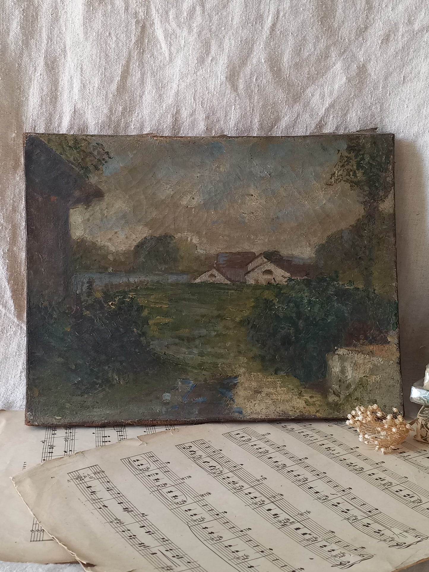 An antique oil landscape painting on canvas depicting a quiet village scene, styled on top of vintage sheet music against a textured white fabric background.