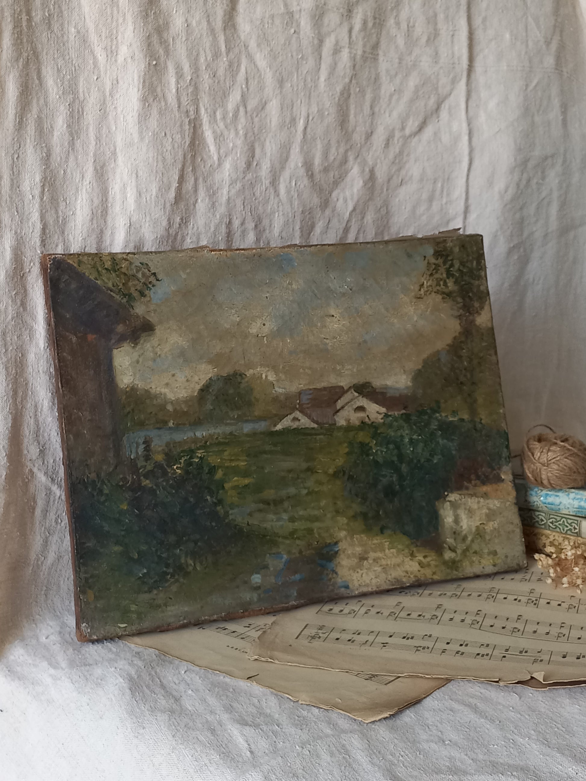 An antique oil landscape painting on canvas, depicting a rural scene with houses, trees, and a river, displayed on vintage sheet music against a draped white linen background.